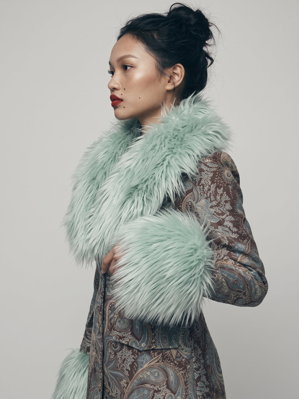 A model in profile wearing a light blue - light mint green shearling   collar and matching fur cuffs over a brown and blue / gray  tone multicolor paisley patterned tapestry coat, against a plain light gray background.