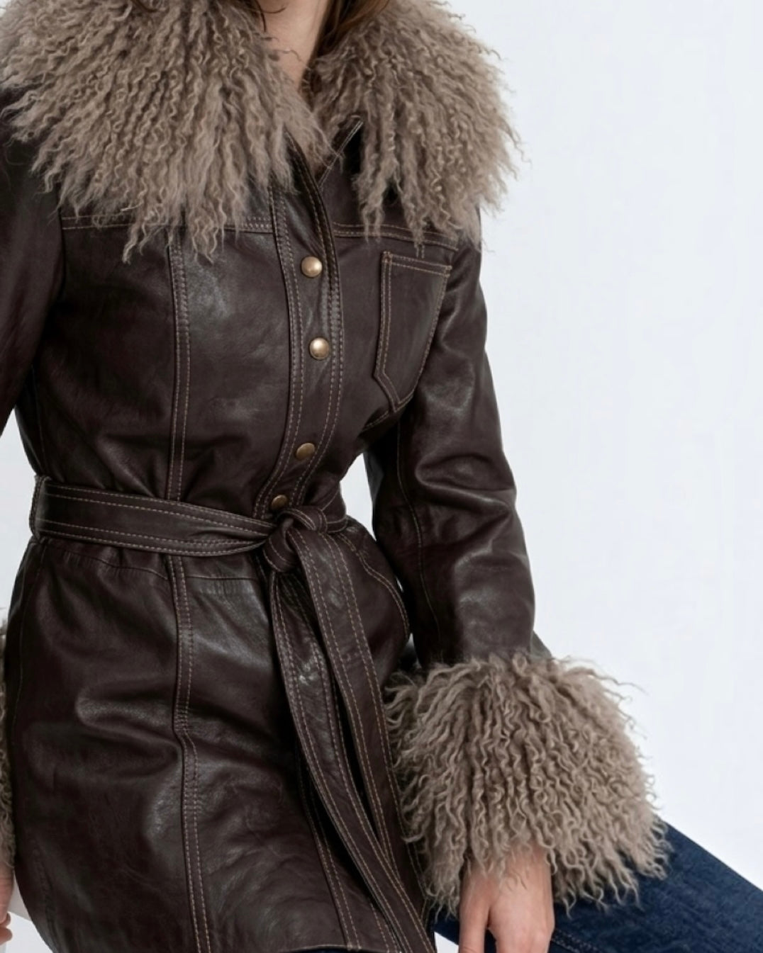 A close-up of a dark brown leather belted coat with a large shaggy beige Mongolian fur collar and matching fur-trimmed cuffs, worn by a model.