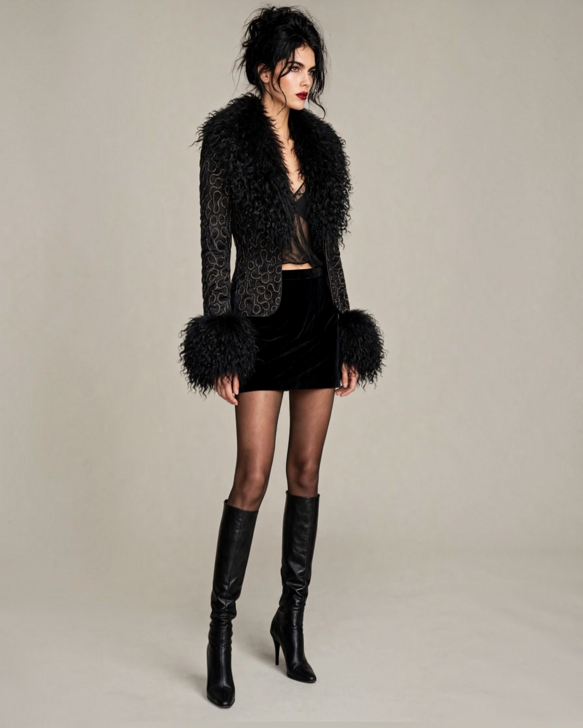 A model wearing a black textured jacket with fluffy Mongolian curly crimped shaggy real lamb fur trim at the collar and cuffs, a black body-con velvet mini skirt, sheer black tights, and tall skinny black leather knee-high high heel boots, standing 3/4  pose against a neutral white  studio background with hair in a tousled updo dark brunette , olive complexion, smoky eye makeup and red lipstick. Sheer black lingerie tank top is worn under the jacket , and her black bra shows subtly . 